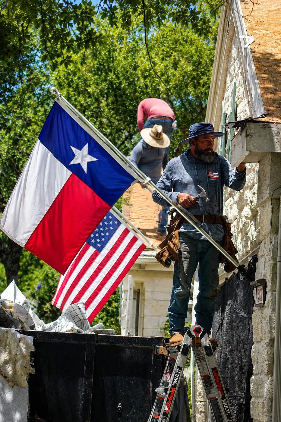 Roman, Benny's brother, on a ladder replacing fascia and siding detail on an Austin home, with Texas and US flags flying